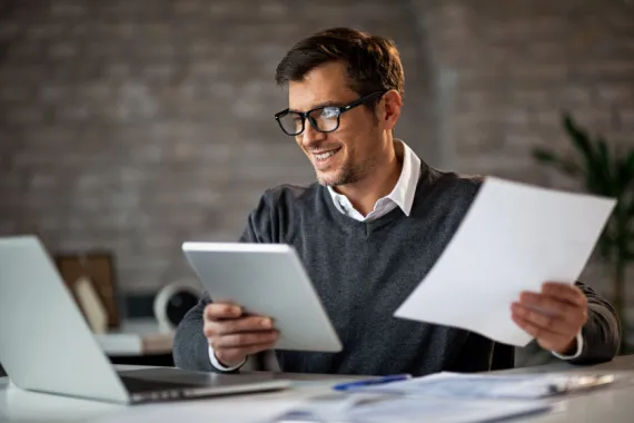 happy businessman using touchpad while looking at a laptop and holding a piece of paper in office