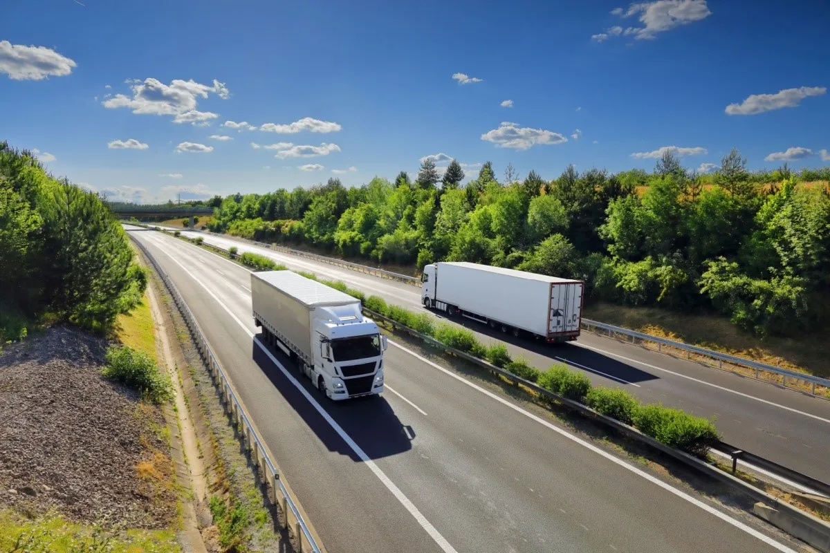 two cargo trucks driving on freeway going opposite directions with a blue sky and green trees on both sides