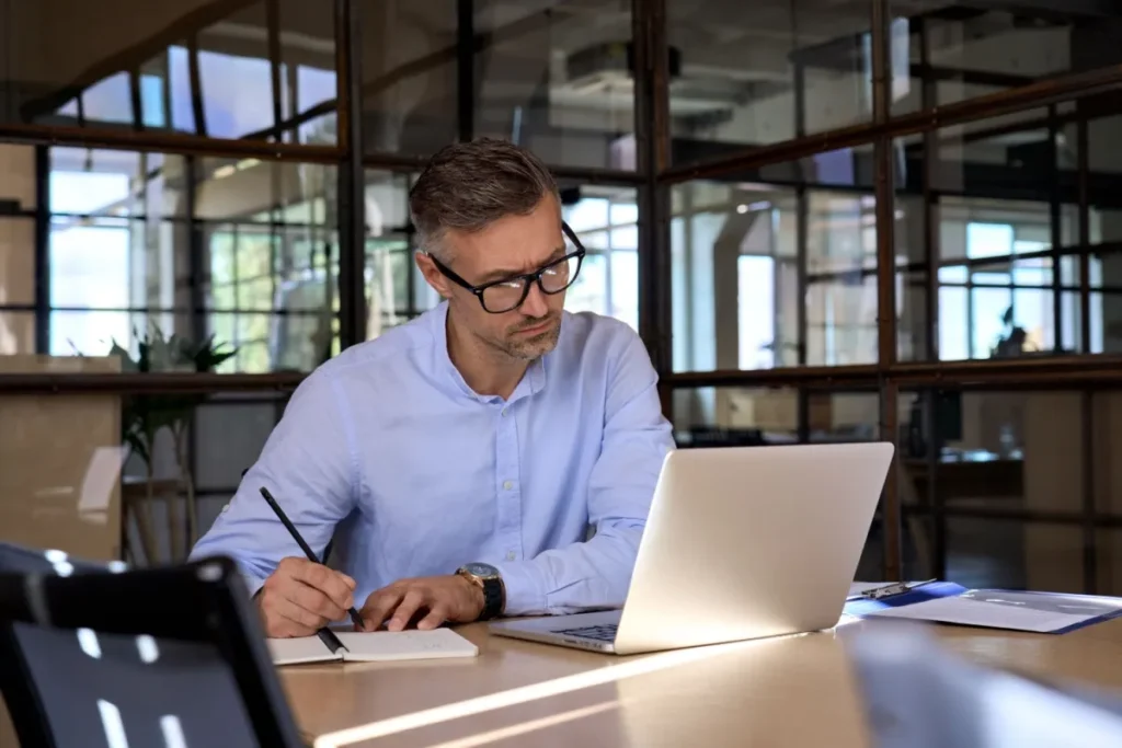 executive business man sitting at office desk looking at laptop while writing things down on notepad